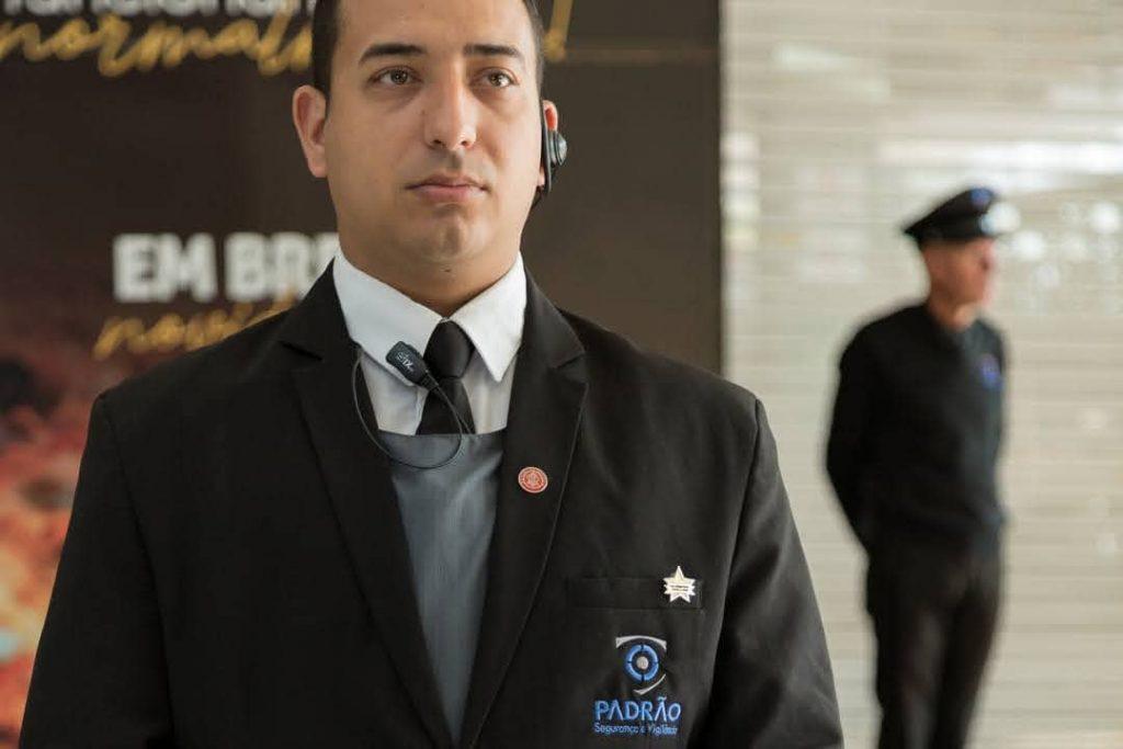 Vigilante uniformizado em destaque, usando terno preto e fone de comunicação, transmitindo profissionalismo e segurança.