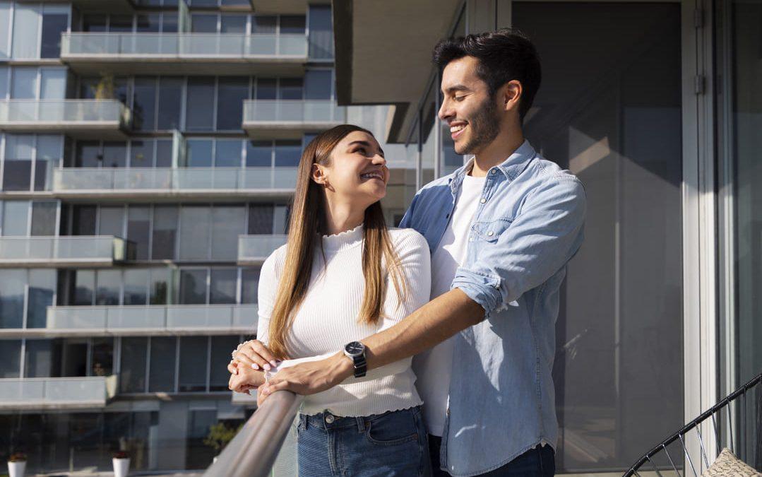 Casal sorridente em sacada de apartamento moderno, com fachada envidraçada ao fundo.