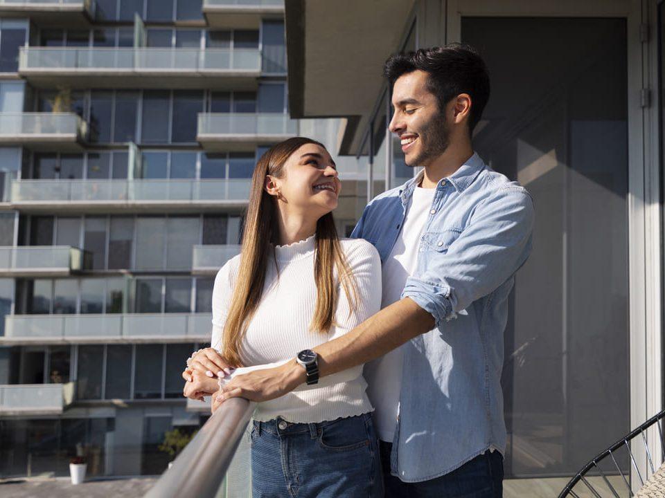 Casal sorridente em sacada de apartamento moderno, com fachada envidraçada ao fundo.