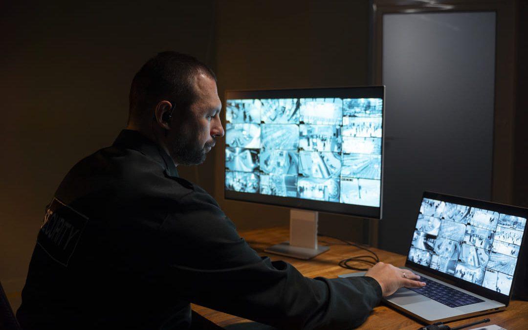 Um segurança monitorando câmeras de vigilância em duas telas, uma de computador e outra de laptop, em um ambiente escuro.