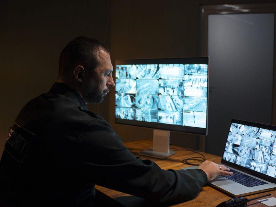 Um segurança monitorando câmeras de vigilância em duas telas, uma de computador e outra de laptop, em um ambiente escuro.