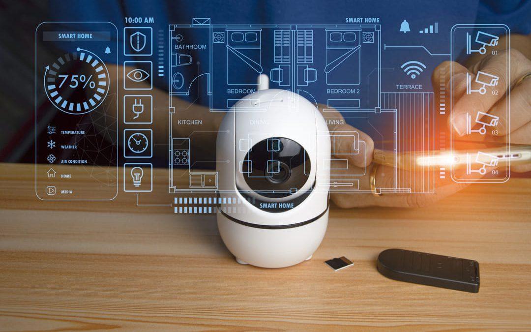 Um conceito de casa inteligente apresentado com um dispositivo de segurança, uma câmera IP branca, em destaque sobre uma mesa de madeira clara. Elementos digitais como planta de uma casa e ícones de funcionalidades conectadas (controle de luz, temperatura, câmeras e dispositivos) aparecem sobrepostos à imagem.