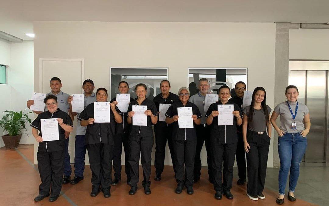 Um grupo de pessoas posando para uma foto em ambiente interno. Eles estão segurando certificados, aparentando terem participado de um treinamento ou curso. Duas mulheres à direita, sem certificados, estão sorrindo e posando junto ao grupo.