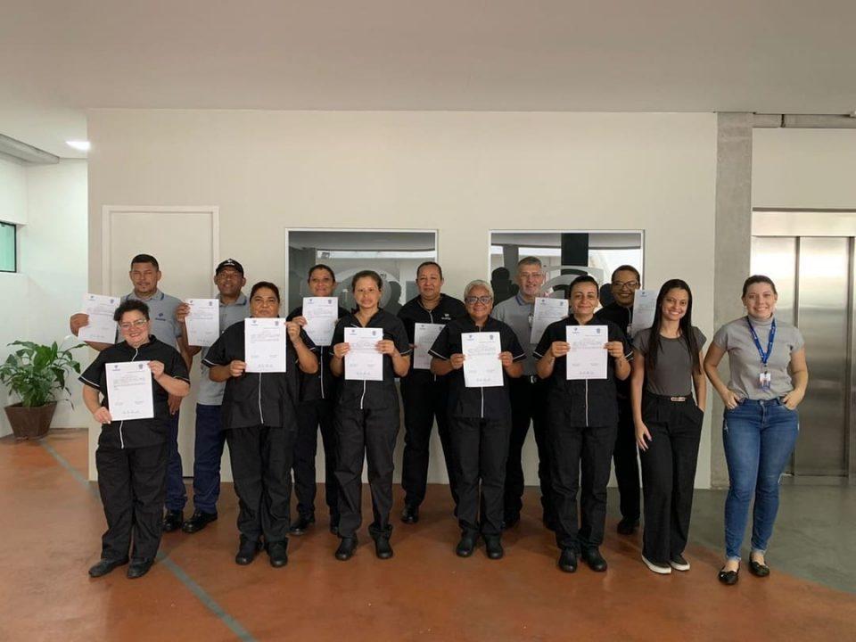 Um grupo de pessoas posando para uma foto em ambiente interno. Eles estão segurando certificados, aparentando terem participado de um treinamento ou curso. Duas mulheres à direita, sem certificados, estão sorrindo e posando junto ao grupo.