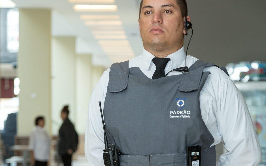 Agente de segurança fardado com colete e rádio comunicador, representando o conceito de segurança preventiva em ambiente corporativo.