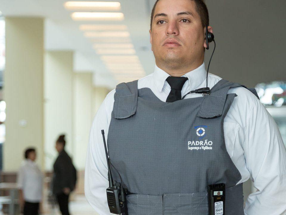Agente de segurança fardado com colete e rádio comunicador, representando o conceito de segurança preventiva em ambiente corporativo.
