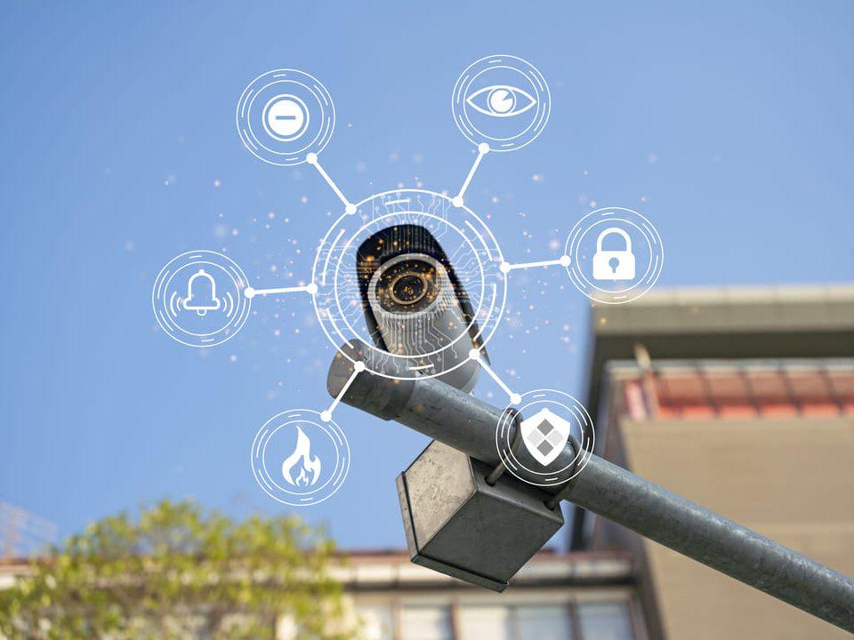 Câmera de segurança moderna com ícones digitais representando inteligência artificial e recursos de segurança, sobre fundo urbano e céu azul.