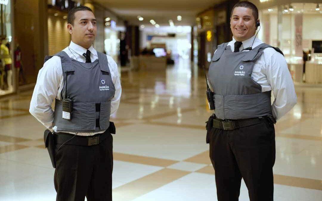 Dois seguranças uniformizados com coletes e rádios em patrulha em um shopping.