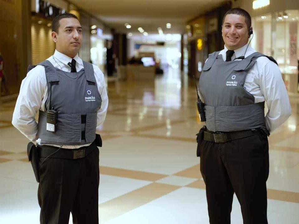 Dois seguranças uniformizados com coletes e rádios em patrulha em um shopping.