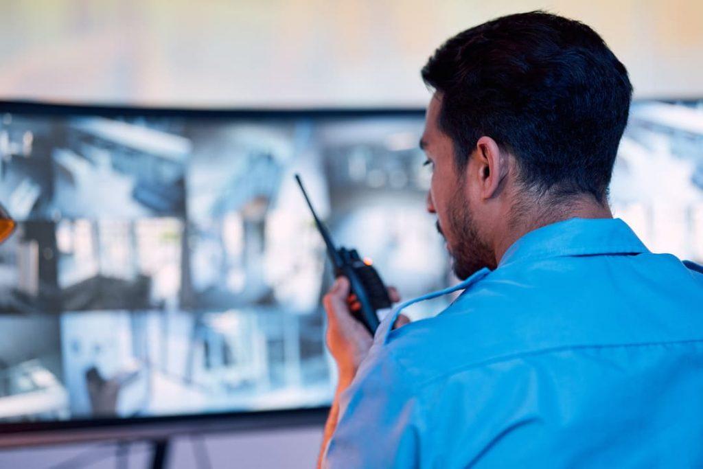 Profissional de segurança monitorando câmeras e se comunicando via rádio em central de portaria remota.