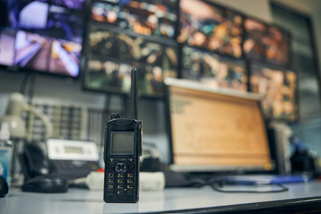 Central de monitoramento com rádio comunicador e múltiplas telas exibindo imagens de câmeras de segurança.