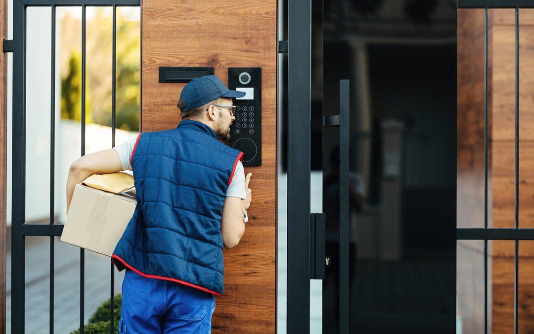 Entregador de uniforme azul segurando uma caixa e falando pelo interfone de um portão moderno de madeira e ferro.