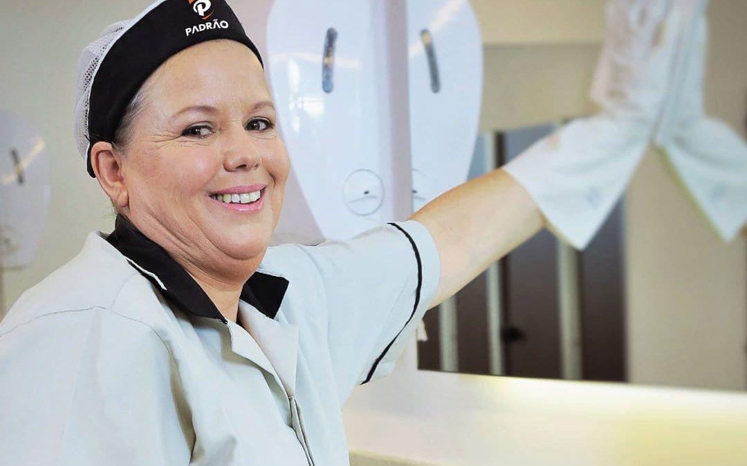 Profissional de limpeza sorridente, usando uniforme branco e touca, higienizando uma superfície com pano.