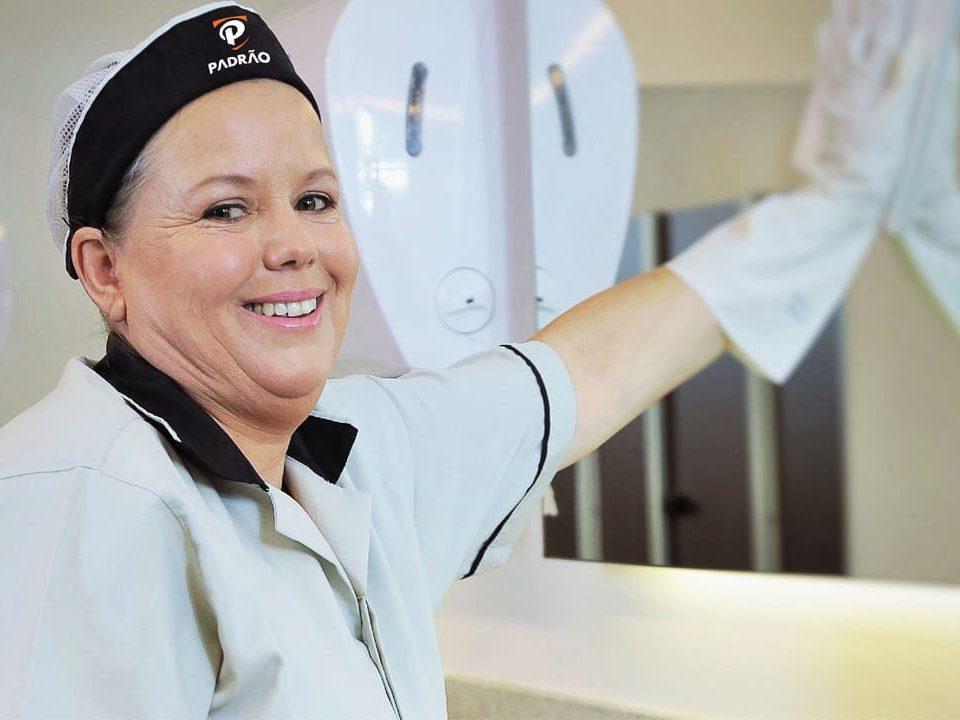 Profissional de limpeza sorridente, usando uniforme branco e touca, higienizando uma superfície com pano.