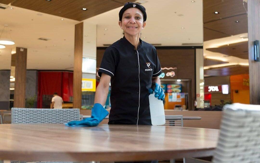 Mulher com uniforme de limpeza e luvas azuis sorri enquanto higieniza uma mesa em praça de alimentação, representando a inclusão social no mercado de trabalho.