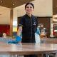 Mulher com uniforme de limpeza e luvas azuis sorri enquanto higieniza uma mesa em praça de alimentação, representando a inclusão social no mercado de trabalho.