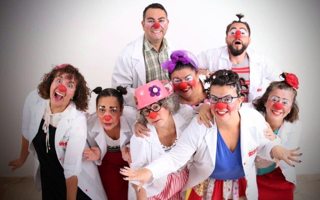Grupo de oito palhaços voluntários vestidos com jalecos brancos, roupas coloridas e narizes vermelhos, posando sorridentes e divertidos para a foto em um ambiente iluminado.