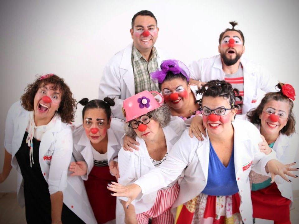 Grupo de oito palhaços voluntários vestidos com jalecos brancos, roupas coloridas e narizes vermelhos, posando sorridentes e divertidos para a foto em um ambiente iluminado.