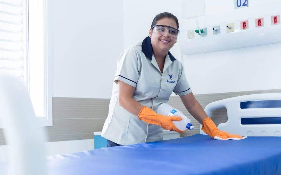 Profissional de limpeza utiliza luvas de proteção enquanto higieniza leito hospitalar com produtos de limpeza.