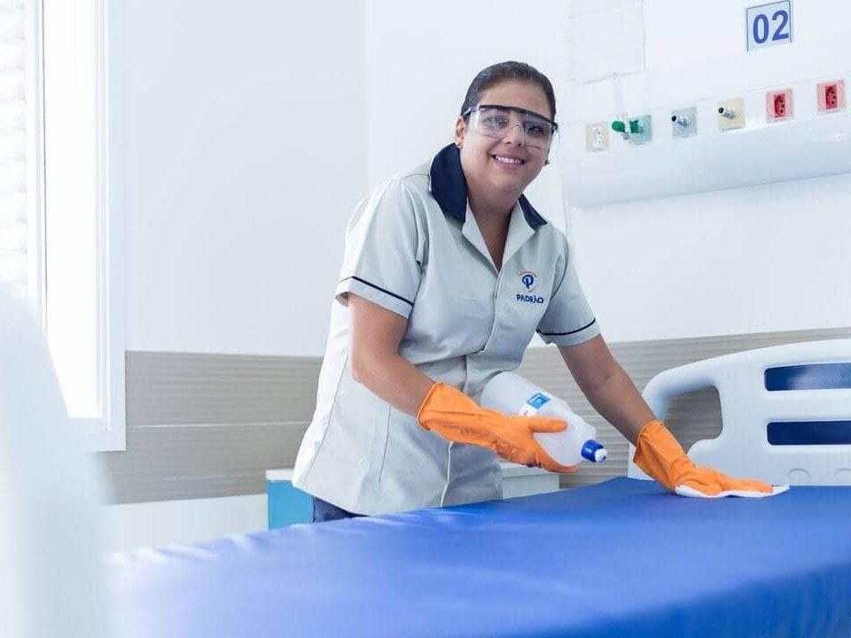 Profissional de limpeza utiliza luvas de proteção enquanto higieniza leito hospitalar com produtos de limpeza.