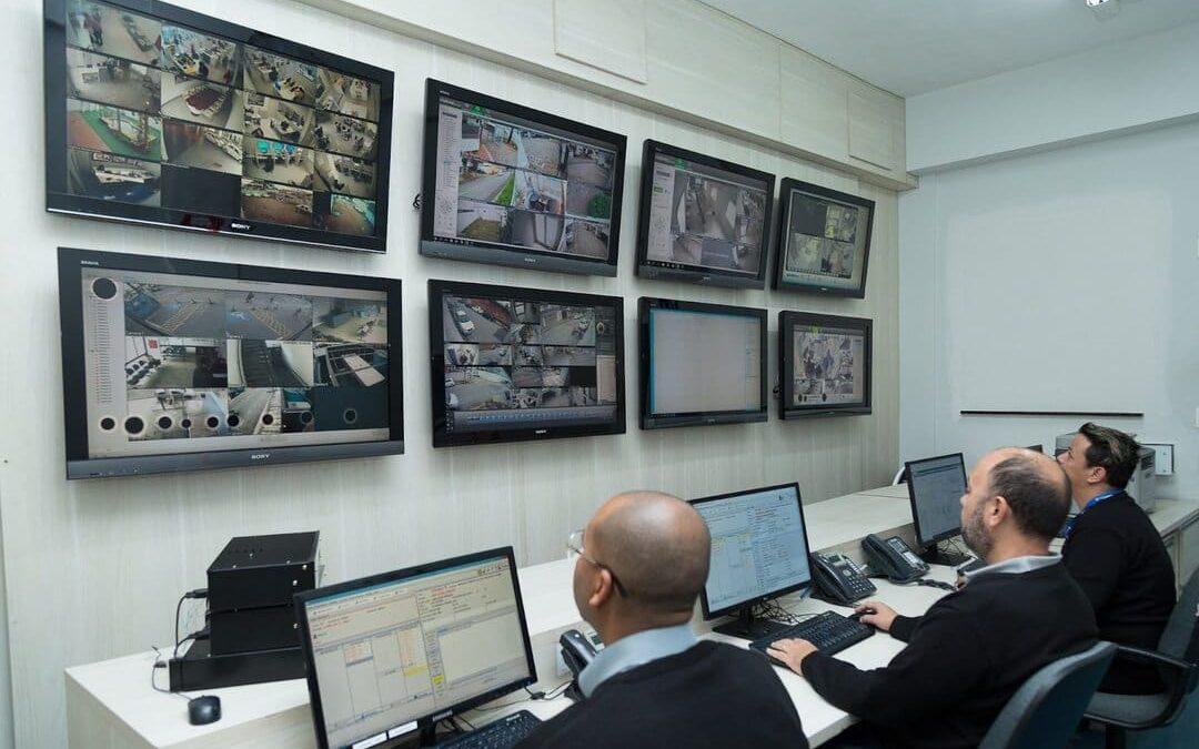 Sala de monitoramento com três pessoas acompanhando câmeras de segurança em vários monitores, representando a gestão condominial e o controle remoto de acesso.