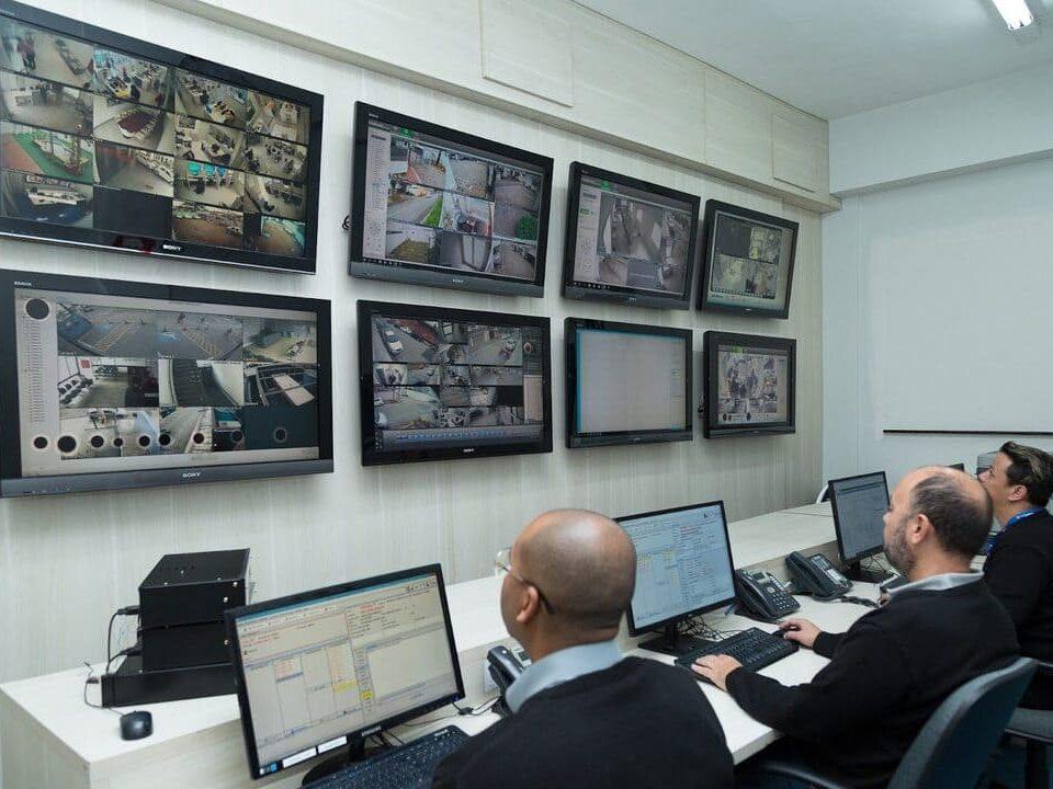 Sala de monitoramento com três pessoas acompanhando câmeras de segurança em vários monitores, representando a gestão condominial e o controle remoto de acesso.