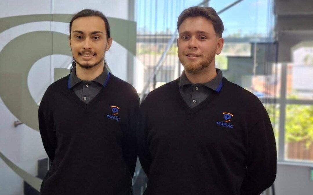 Dois jovens aprendizes uniformizados da empresa Padrão posam sorrindo em um ambiente corporativo moderno e iluminado.