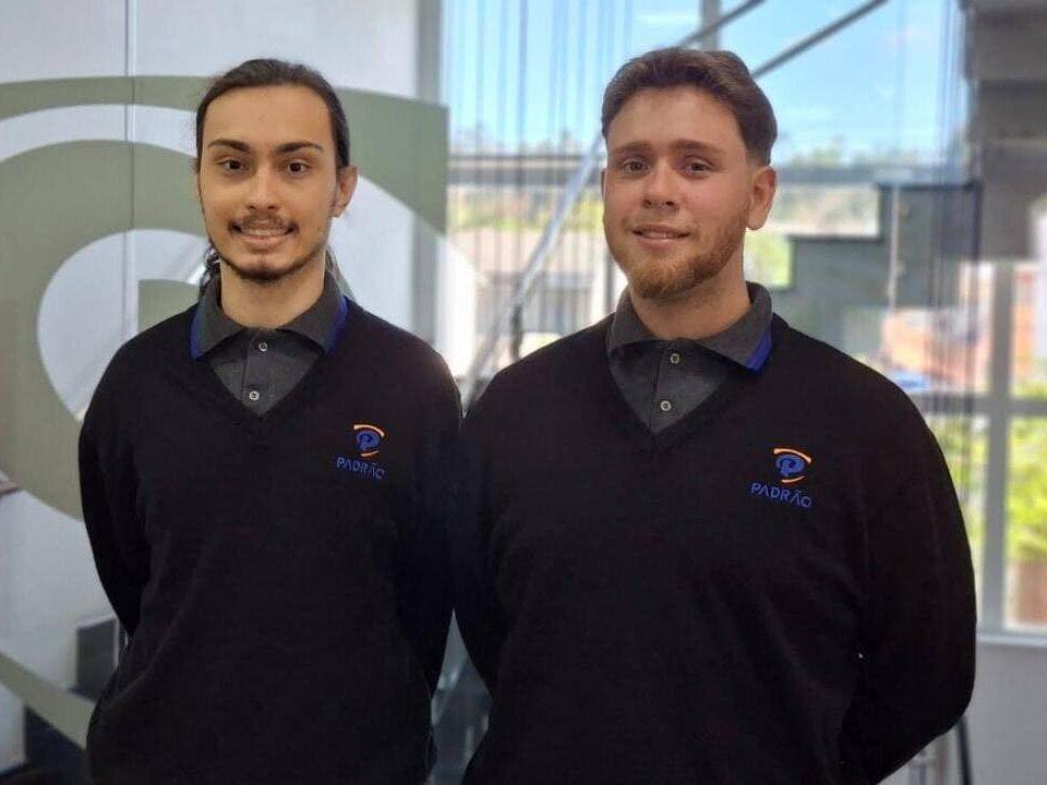 Dois jovens aprendizes uniformizados da empresa Padrão posam sorrindo em um ambiente corporativo moderno e iluminado.