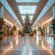 Interior de um shopping center decorado para o Natal, com uma grande árvore iluminada no centro, guirlandas nas colunas e iluminação festiva ao longo dos corredores.