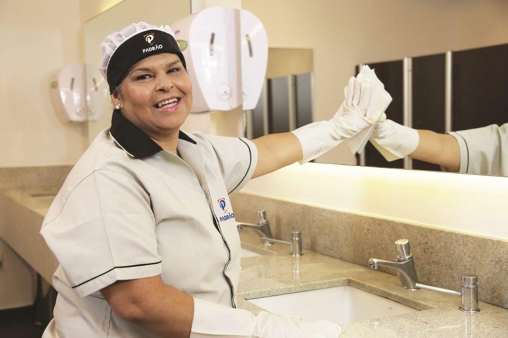 Colaboradora uniformizada realizando a limpeza de um espelho em um banheiro de shopping, utilizando luvas e pano de higienização, enquanto sorri para a câmera.