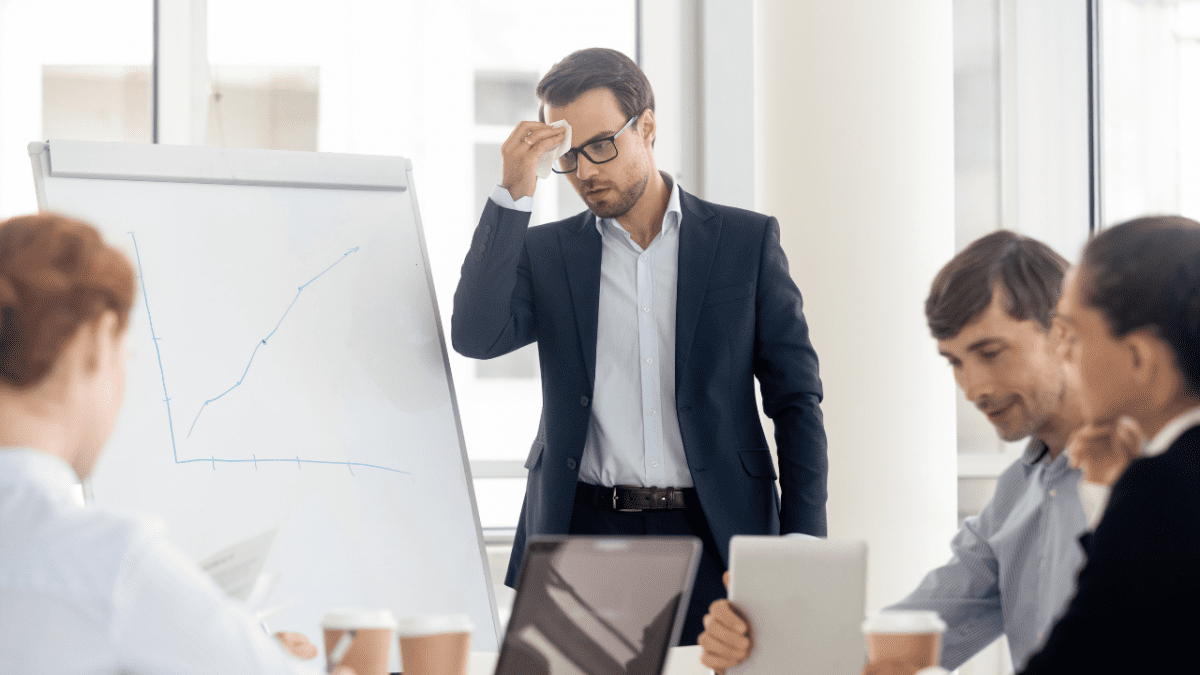 Homem de terno, com expressão preocupada, enxuga a testa diante de um quadro com gráfico enquanto colegas trabalham ao redor.