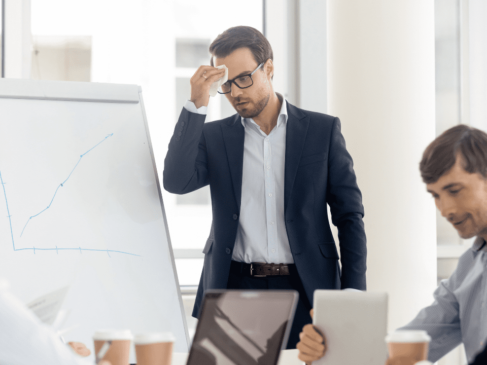Homem de terno, com expressão preocupada, enxuga a testa diante de um quadro com gráfico enquanto colegas trabalham ao redor.