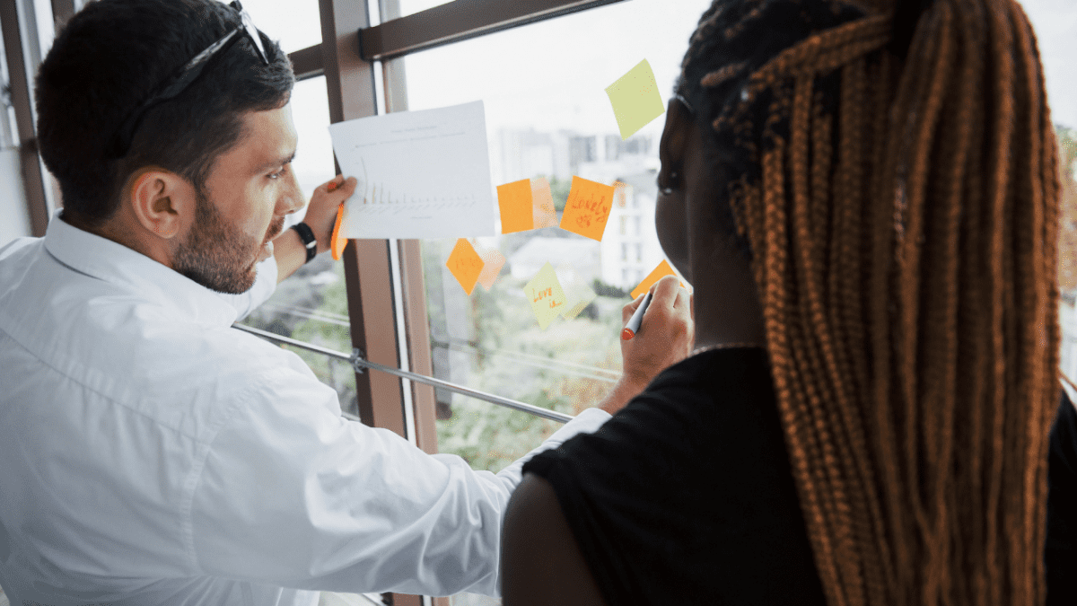 Duas pessoas analisam gráfico e escrevem em post-its colados em vidro.