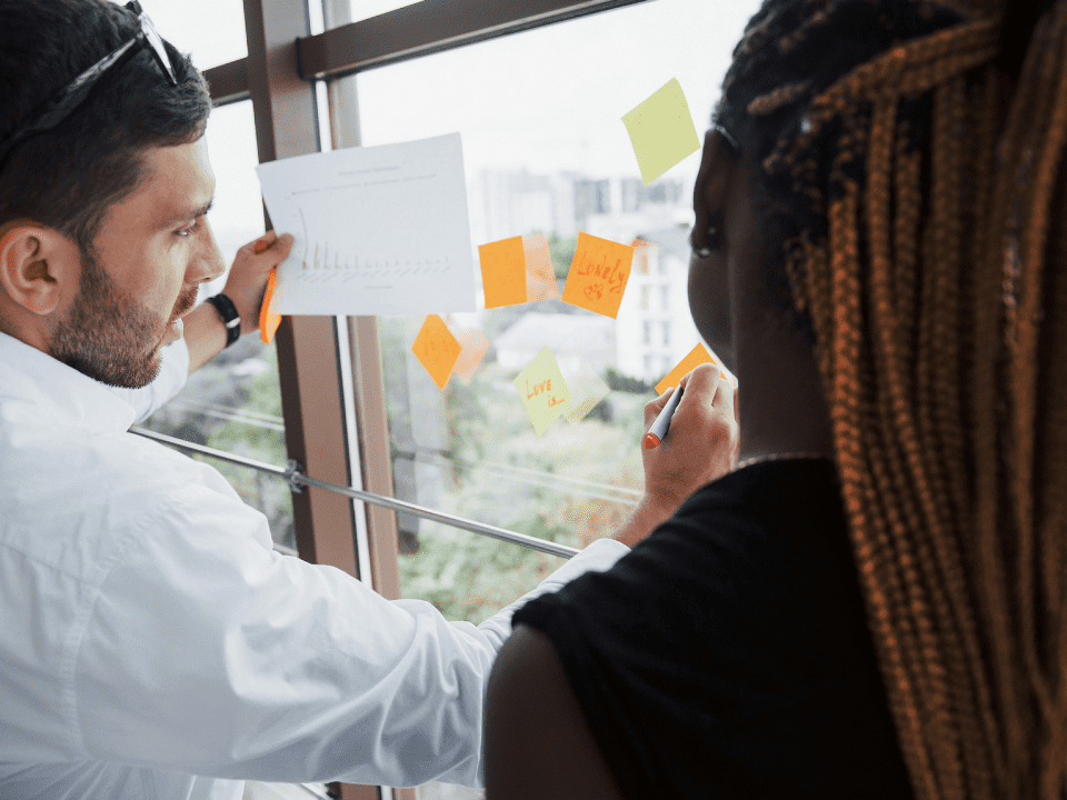 Duas pessoas analisam gráfico e escrevem em post-its colados em vidro.