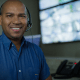 Profissional de monitoramento usando headset em central de segurança com telas de vigilância ao fundo.