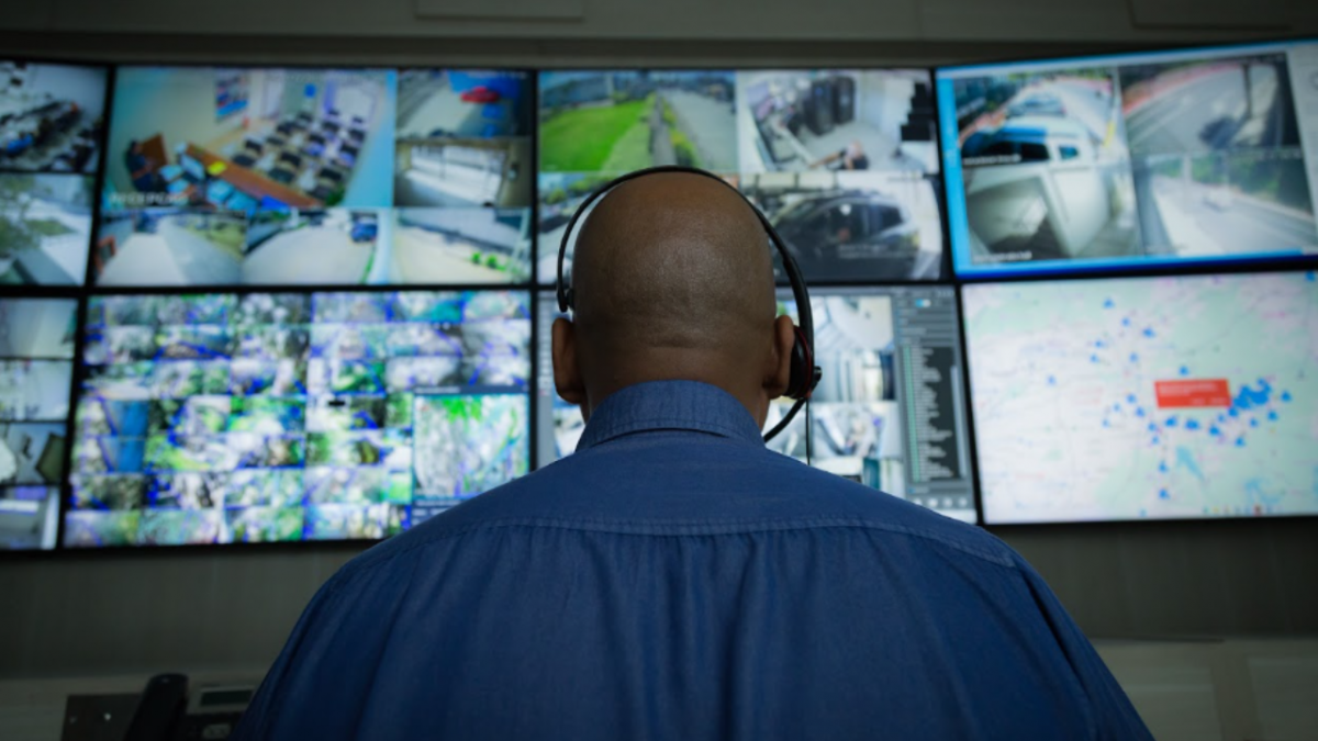 Operador monitora câmeras de segurança em uma central de vigilância com múltiplos monitores.