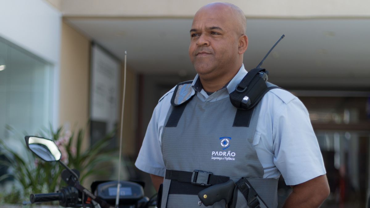 Vigilante uniformizado em frente a um edifício, utilizando rádio comunicador e colete de segurança.