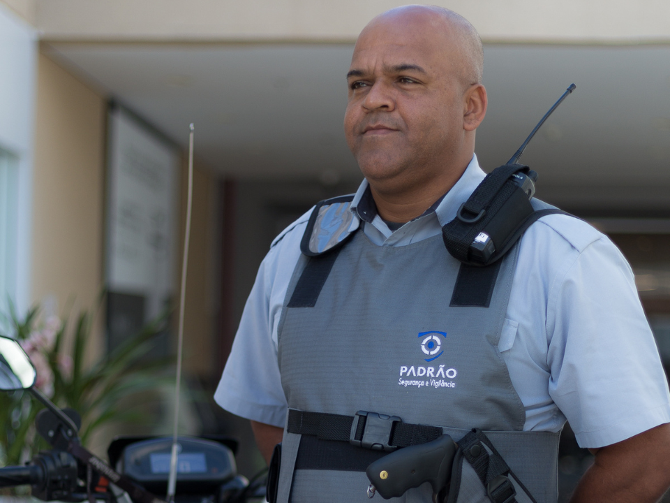 Vigilante uniformizado em frente a um edifício, utilizando rádio comunicador e colete de segurança.