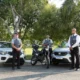 Equipe de segurança em condomínio posicionada ao lado de dois carros e uma motocicleta de patrulhamento, em uma área arborizada.
