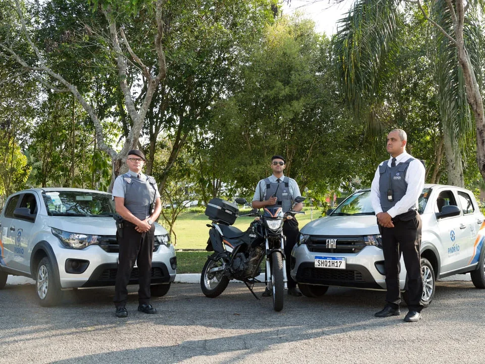 Equipe de segurança em condomínio posicionada ao lado de dois carros e uma motocicleta de patrulhamento, em uma área arborizada.