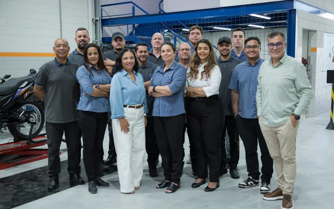 Um grupo diversificado de funcionários de uma oficina de motocicletas posa para uma foto de grupo, simbolizando o fim de um treinamento de funcionário. Eles estão sorrindo para a câmera, vestidos com o uniforme da empresa. Ao fundo, uma motocicleta em um elevador de manutenção e uma área de mezanino.
