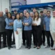 Um grupo diversificado de funcionários de uma oficina de motocicletas posa para uma foto de grupo, simbolizando o fim de um treinamento de funcionário. Eles estão sorrindo para a câmera, vestidos com o uniforme da empresa. Ao fundo, uma motocicleta em um elevador de manutenção e uma área de mezanino.