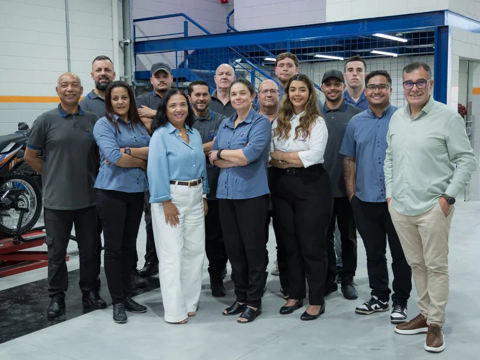 Um grupo diversificado de funcionários de uma oficina de motocicletas posa para uma foto de grupo, simbolizando o fim de um treinamento de funcionário. Eles estão sorrindo para a câmera, vestidos com o uniforme da empresa. Ao fundo, uma motocicleta em um elevador de manutenção e uma área de mezanino.